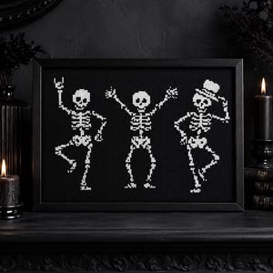 May include: A framed cross-stitch embroidery featuring three white skeletons on a black background. The left skeleton makes a rock-and-roll hand gesture, the center skeleton has arms raised, and the right skeleton wears a top hat while dancing.