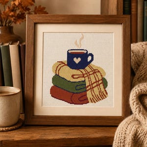 May include: A framed cross-stitch embroidery featuring a stack of three folded sweaters in yellow, green, and dark red, topped with a blue mug decorated with a white heart and rising steam, all set against a white fabric background.