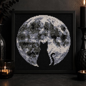 May include: A square framed cross-stitch embroidery featuring a solid black silhouette of a cat sitting in front of a large, detailed full moon. The moon is rendered in shades of white, grey, and dark blue, set against a solid black background.