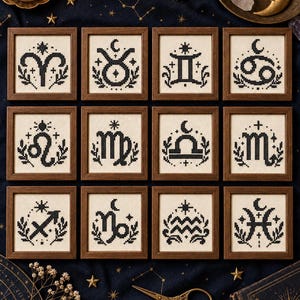 May include: Twelve square wooden frames containing black cross-stitch embroidery on cream fabric, each depicting a unique zodiac sign symbol surrounded by stars, moons, and foliage motifs, arranged in a grid pattern on a dark blue fabric surface.