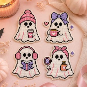 May include: Four cross-stitch ghost patches featuring white ghosts with black eyes. One wears a pink beanie holding a cup, one has a purple bow holding a pink mug, one wears pink earmuffs reading a purple book, and one wears a pink headband holding a mirror and bottle.