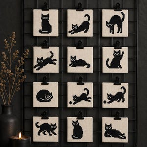 May include: A black metal grid wall display holds twelve square fabric pieces, each featuring a unique black cross-stitch cat silhouette on a cream background. The cats are depicted in various poses, including sitting, stretching, playing, and lying down.