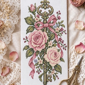 May include: A rectangular cross-stitch embroidery on white fabric featuring a central ornate key in shades of green and gold, adorned with two large pink roses, smaller floral accents, and a flowing pink ribbon tied in a bow at the top.