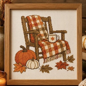 May include: A framed cross-stitch embroidery featuring a wooden chair draped with an orange and white plaid blanket, a mug with a small leaf design, two pumpkins, and scattered autumn leaves on a cream-coloured fabric background.