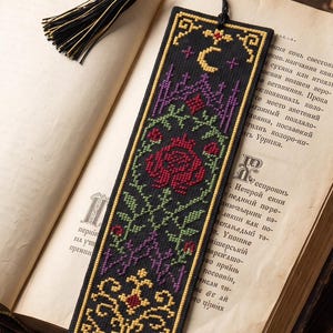 Può includere: Un segnalibro rettangolare in tessuto nero con ricami a punto croce nei colori rosso, verde, viola e giallo. Il design presenta una luna crescente, motivi floreali e bordi geometrici, completato da una nappa nera e oro sulla parte superiore.