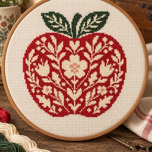 May include: A circular wooden embroidery hoop containing a cross-stitch pattern of a red apple on white fabric. The apple features intricate white floral and leaf motifs within its shape, topped with two dark green leaves. The design is symmetrical.