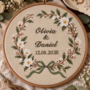 Op de afbeelding: Een ronde houten borduurring met beige stof en een in kruissteek geborduurde bloemenkrans in groen, wit en bruin. In het midden staat de tekst Olivia & Daniel en de datum 12.06.2026 in donkergroene draad, afgewerkt met een strik.