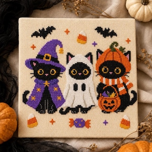May include: A square cross-stitch embroidery on beige fabric featuring three black cats in Halloween costumes: one as a purple-clad witch, one as a white ghost, and one wearing an orange pumpkin suit while holding a jack-o'-lantern candy bucket.