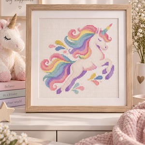 Puede incluir: Un bordado en punto de cruz cuadrado que muestra un unicornio blanco con una melena y cola de colores del arcoíris, enmarcado en un marco de madera de tono claro con un paspartú blanco. El unicornio está representado de perfil.