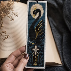 May include: A rectangular cross-stitch bookmark featuring a black swan with a gold crown against a cream-coloured moon and a dark blue background. The lower section displays a white fleur-de-lis emblem with gold accents and a black cord at the top.