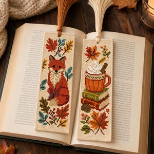 May include: Two rectangular cross-stitch bookmarks made of cream-coloured fabric. The left bookmark features a red fox surrounded by colourful autumn leaves. The right bookmark displays a mug of hot cocoa on books with leaves. Both have tassels.