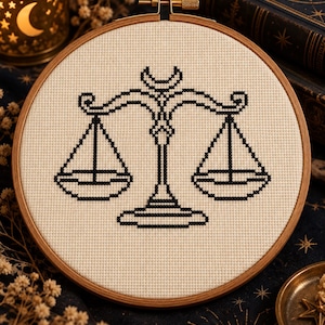 May include: A circular wooden embroidery hoop containing a piece of off-white fabric with a black cross-stitch design of a balance scale. The scale features a central pillar topped with a crescent moon shape and two hanging pans on either side.