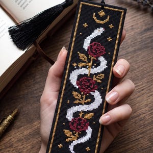 May include: A rectangular black fabric bookmark featuring a cross-stitched design of a white snake winding through three deep red roses. The embroidery includes gold-colored stars and a crescent moon at the top, finished with a black tassel.