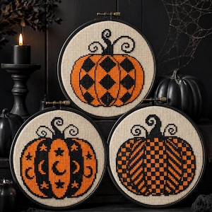 Peut inclure: Trois tambours à broder circulaires présentant des motifs de citrouilles au point de croix sur tissu crème. Une citrouille a un motif losange noir et orange, une autre des motifs de lune et d'étoiles, et la troisième un damier noir et orange.