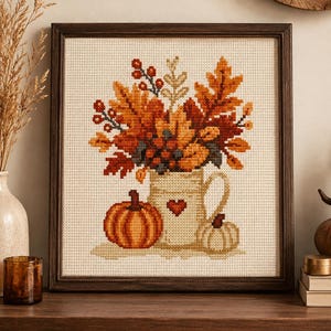 Könnte beinhalten: Ein gerahmtes Kreuzstich-Stickbild mit einem cremefarbenen Krug, der mit Herbstblättern, Beeren und Laub gefüllt ist. Links neben dem Krug befindet sich ein leuchtend orangefarbener Kürbis, während rechts ein kleinerer, blasser Kürbis steht.