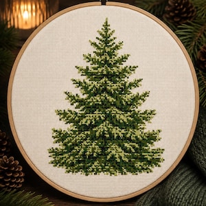 May include: A circular wooden embroidery hoop containing a cross-stitch design of a coniferous tree. The tree is rendered in various shades of green thread against a plain white fabric background, featuring detailed needlework texture.