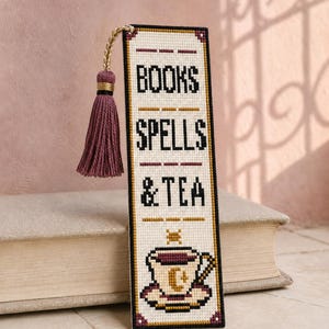Puede incluir: Un marcapáginas rectangular de punto de cruz con el texto negro BOOKS SPELLS & TEA sobre un gráfico de una taza de té con un diseño de media luna. El marcapáginas tiene un fondo color crema, un borde negro y una borla malva en la parte superior.
