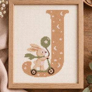 May include: A framed cross-stitch embroidery featuring a large tan letter J decorated with white stars and crescent moons. A small tan rabbit with a green scarf rides a green scooter while holding a green balloon, all on white fabric.