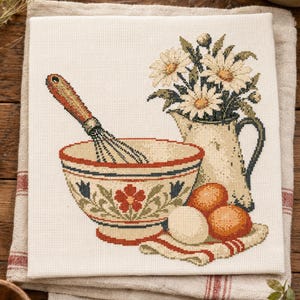 Peut inclure: Une broderie au point de croix sur tissu blanc représentant une scène de cuisine avec un bol à mélanger, un fouet, une cruche remplie de marguerites blanches et trois œufs posés sur un petit linge à rayures rouges, dans des tons terreux.