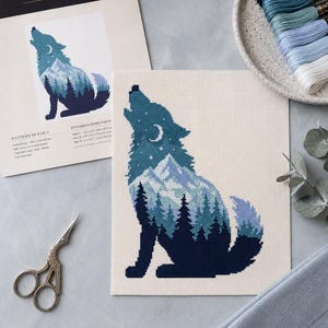 Może przedstawiać: Wz&oacute;r do haftu krzyżykowego zatytułowany Howling Wolf przedstawiający sylwetkę wilka wypełnioną g&oacute;rskim i leśnym krajobrazem w odcieniach niebieskiego, bieli i granatu. Zawiera drukowaną instrukcję, nici do haftowania oraz metalowe nożyczki.