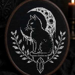 May include: A cross-stitch embroidery piece in a dark wooden oval hoop featuring a white thread design on black fabric. The pattern depicts a cat sitting in front of a crescent moon, framed by two leafy branches with a crystal hanging at the bottom.