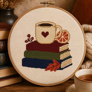Peut inclure: Un tambour à broder circulaire en bois présentant un motif au point de croix d'une tasse crème avec un cœur foncé, posée sur une pile de trois livres bordeaux, verts et bleus. Une tranche d'orange, des baies rouges et une feuille sont inclus.