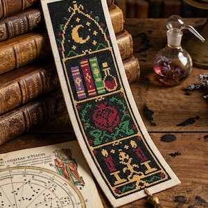 May include: A rectangular fabric bookmark featuring intricate cross-stitch embroidery on a black background. The design includes a crescent moon, colorful books, a potion bottle, a red flower, and candles, finished with a gold-colored tassel.