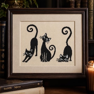 May include: A framed cross-stitch embroidery featuring three black cats on a beige fabric background. The cats are depicted in different poses: one stretching, one sitting upright, and one crouching low, all with long, curled tails.