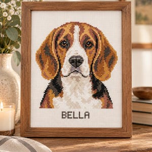 May include: A framed cross-stitch portrait of a beagle dog with brown, black, and white fur. The dog is centred on a white fabric background with the name BELLA stitched in black capital letters underneath the dog's chin, all inside a wooden frame.