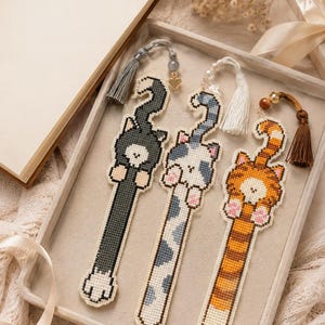 Puede incluir: Tres marcapáginas de punto de cruz con forma de gato, cada uno con un patrón de pelaje diferente: gris oscuro sólido, blanco con manchas grises y atigrado naranja. Cada marcapáginas incluye una borla decorativa y cuentas en el extremo superior.