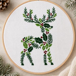 May include: A circular wooden embroidery hoop containing white fabric with a cross-stitch design of a reindeer. The reindeer is composed of green holly leaves, pine branches, and small red berries, creating a festive botanical silhouette.