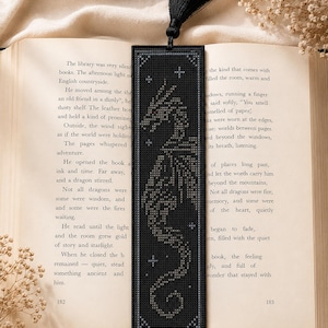 Peut inclure: Un marque-page rectangulaire noir orné d'un motif au point de croix représentant un dragon à l'œil rouge, entouré de petites étoiles blanches. Le marque-page est surmonté d'un pompon noir et posé verticalement sur une page de livre ouverte.