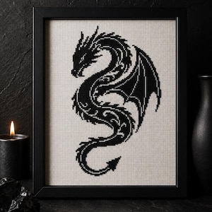 以下が含まれることがあります： 薄いベージュ色のアイーダ布に黒いドラゴンのシルエットが刺繍された、クロスステッチの額入り作品。ドラゴンは横向きの姿で、体、翼、尾に複雑な模様が施されており、シンプルな黒いフレームに収められています。
