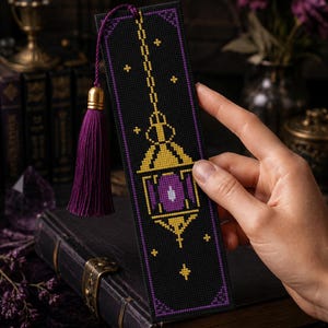 Può includere: Un segnalibro rettangolare nero con un disegno a punto croce di una lanterna dorata con un centro viola. Il segnalibro è bordato da una linea viola e include una nappa viola fissata sulla parte superiore con un cappuccio in metallo dorato.