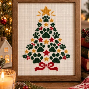 Könnte beinhalten: Ein gerahmtes Kreuzstichbild auf weißem Stoff, das einen Weihnachtsbaum aus dunkelgrünen Pfotenabdrücken, gelben Sternen und kleinen roten Stechpalmenbeeren zeigt. Der Baum wird von einem großen gelben Stern und einer roten Schleife gekrönt.