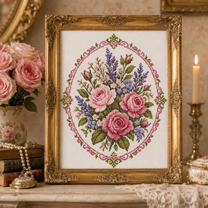 Puede incluir: Un bordado de punto de cruz que presenta un ramo de rosas rosas, lavanda morada y pequeñas flores azules, encerrado dentro de un borde ovalado decorativo de patrones geométricos rosas y verdes, todo en un marco rectangular dorado ornamentado.