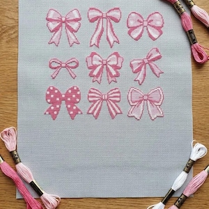 May include: A light grey cross-stitch fabric featuring a grid of nine distinct pink bows in various patterns including polka dots and stripes, accompanied by several loose skeins of pink and white embroidery thread.