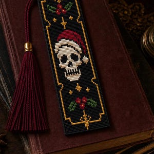 Puede incluir: Un marcapáginas rectangular de punto de cruz con fondo negro y una calavera blanca central con un gorro de Papá Noel rojo. El diseño incluye bordes decorativos dorados, hojas de acebo verdes y bayas rojas, terminado con una borla roja.