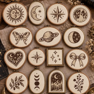 May include: A collection of fourteen small embroidery hoops and frames featuring dark brown cross-stitch designs on cream fabric. Motifs include a sun, moon, stars, planet, butterfly, heart, bow, botanical sprigs, and a mountain landscape scene.
