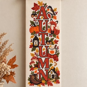 Puede incluir: Un tapiz vertical de punto de cruz con la palabra AUTUMN en letras rojas grandes. El diseño incluye hojas de otoño coloridas, un zorro, una calabaza, una linterna, una taza con bebida, setas, una casa pequeña y botas sobre tela crema.