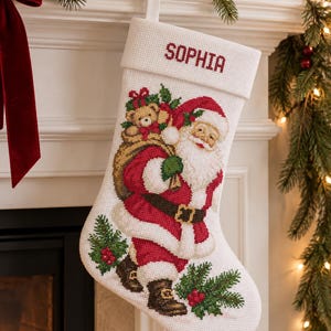 May include: A white Christmas stocking featuring a cross-stitch design of Santa Claus carrying a sack with a teddy bear. The name SOPHIA is embroidered in red on the cuff. The stocking includes green pine sprigs with red berries at the base.