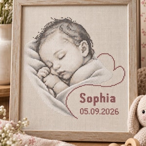 May include: A framed cross-stitch portrait of a sleeping infant wrapped in a blanket. The design features the name Sophia and the date 05.09.2026 stitched in dark red thread. The artwork is encased in a light-toned wooden frame with a square profile.