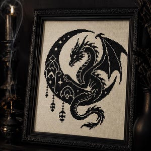 May include: A framed cross-stitch embroidery featuring a black dragon silhouette centred within a crescent moon. The design includes intricate geometric patterns, stars, and hanging ornaments, all stitched in black thread on a light beige fabric.
