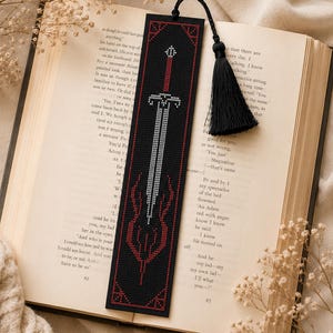 Op de afbeelding: Een rechthoekige zwarte boekenlegger met een kruissteekontwerp van een zilveren zwaard met een rood handvat en rode decoratieve accenten. De boekenlegger is afgewerkt met een zwart koord en een bijpassende zwarte kwast aan de bovenrand.
