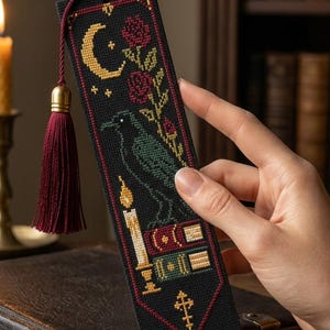 Può includere: Un segnalibro rettangolare in tessuto nero con un ricamo a punto croce raffigurante una mezzaluna gialla, rose rosse, un corvo verde scuro, una candela accesa e libri impilati, rifinito con una nappa di colore rosso scuro in cima.