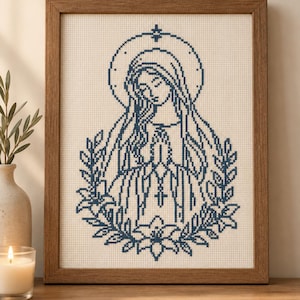 Könnte beinhalten: Ein gerahmtes Kreuzstichbild mit einem dunkelblauen Fadenmotiv einer Figur mit Heiligenschein, die Hände zum Gebet gefaltet, umgeben von einem Kranz aus Blättern und Blumen auf einem hell cremefarbenen Stoffhintergrund.
