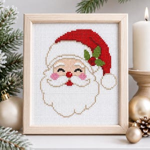 Puede incluir: Un bordado de punto de cruz de Santa Claus en un marco de madera clara. El diseño presenta un gorro rojo con un pompón blanco, hojas de acebo verdes, una nariz roja, mejillas rosadas y una barba blanca sobre tela blanca con cuadrícula.