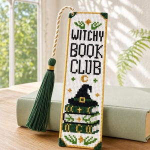 Może przedstawiać: Prostokątna zakładka do książki wykonana haftem krzyżykowym z napisem WITCHY BOOK CLUB czarnymi literami. Wzór przedstawia stos książek zwieńczony czarnym kapeluszem czarownicy, ozdobiony zielonymi i złotymi motywami oraz zielonym chwostem.