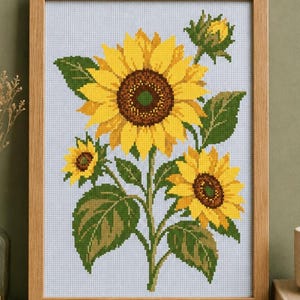 May include: A cross-stitch embroidery of three sunflowers and a bud in a light wooden frame. The design features bright yellow petals, dark brown centres, and green leaves on a light blue grid fabric. The artwork is displayed vertically.