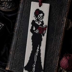 Puede incluir: Un marcapáginas rectangular con un diseño de punto de cruz de una figura esquelética con un vestido largo negro y una flor roja en el pelo. La figura sostiene un libro rojo abierto. El marcapáginas es de tela color hueso con una borla roja oscura.
